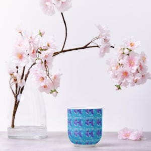 May include: A blue ceramic mug with a repeating pattern of purple fish and bubbles. The mug is set against a backdrop of pink cherry blossom branches in a clear vase. A few pink blossoms are scattered on the surface.