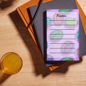 May include: A notepad with the word "Notes" in cursive, featuring a lavender background and colorful, oval-shaped designs. The notepad is lined and rests on a stack of books with a pen and a glass of yellow liquid.