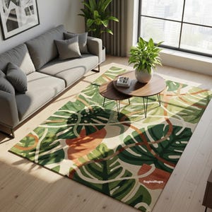 Puede incluir: Una alfombra estampada con un diseño de hojas tropicales en un salón. La alfombra presenta grandes hojas verdes de monstera con acentos naranjas y marrones sobre un fondo beige. La habitación incluye un sofá gris y mesas de centro de madera.