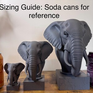May include: Three gray elephant sculptures of varying sizes are displayed next to two soda cans for size comparison. The text "Sizing Guide: Soda cans for reference" is at the top of the image. The sculptures are on a wooden surface.
