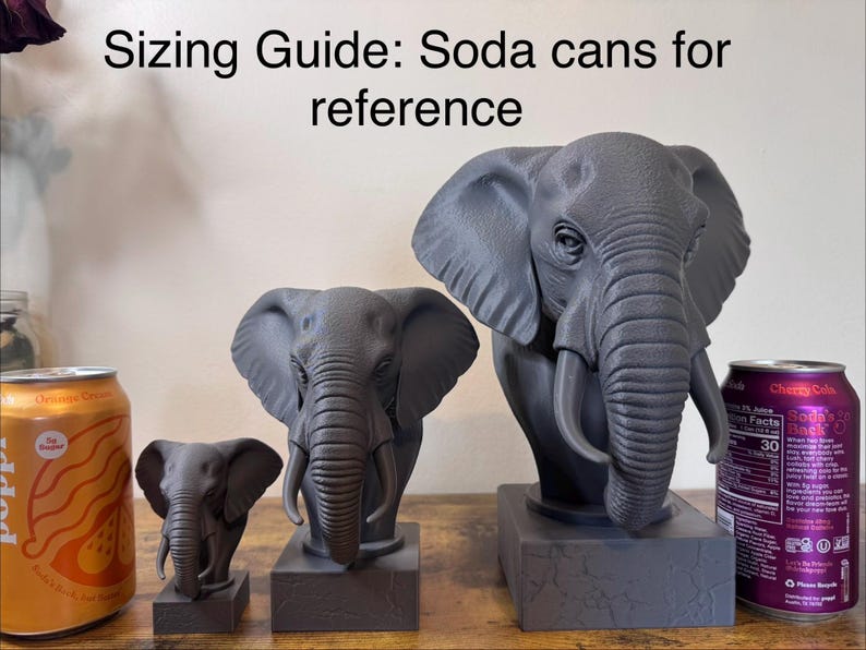 May include: Three gray elephant sculptures of varying sizes are displayed next to two soda cans for size comparison. The text "Sizing Guide: Soda cans for reference" is at the top of the image. The sculptures are on a wooden surface.