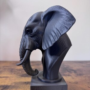 May include: A dark gray elephant bust sculpture. The sculpture features detailed textures and a realistic depiction of an elephant's head, trunk, and ear. The bust is mounted on a rectangular base, creating a striking decorative piece.