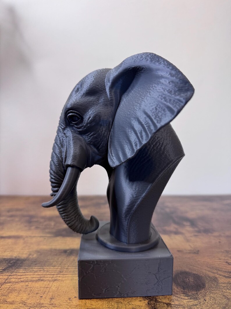 May include: A dark gray elephant bust sculpture. The sculpture features detailed textures and a realistic depiction of an elephant's head, trunk, and ear. The bust is mounted on a rectangular base, creating a striking decorative piece.