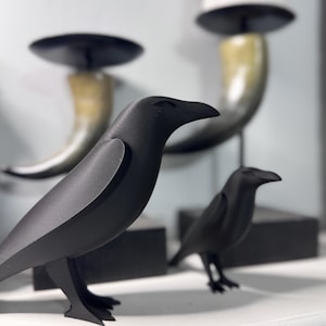 May include: Two black raven figurines with a matte finish are displayed on a white surface. The ravens are in different sizes and poses. In the background, there are two candle holders with black tops and horn-shaped bases.