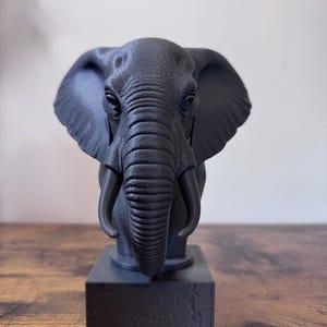 May include: A dark gray elephant bust sculpture with detailed wrinkles and tusks. The elephant is mounted on a square base. The sculpture is a decorative item.