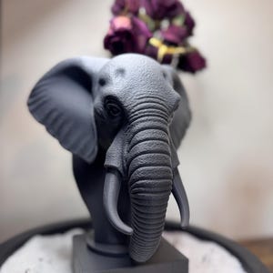 May include: A gray elephant bust with detailed features, including large ears and tusks. The sculpture is mounted on a square base and adorned with a bouquet of deep purple flowers. The overall design is a blend of art and nature.