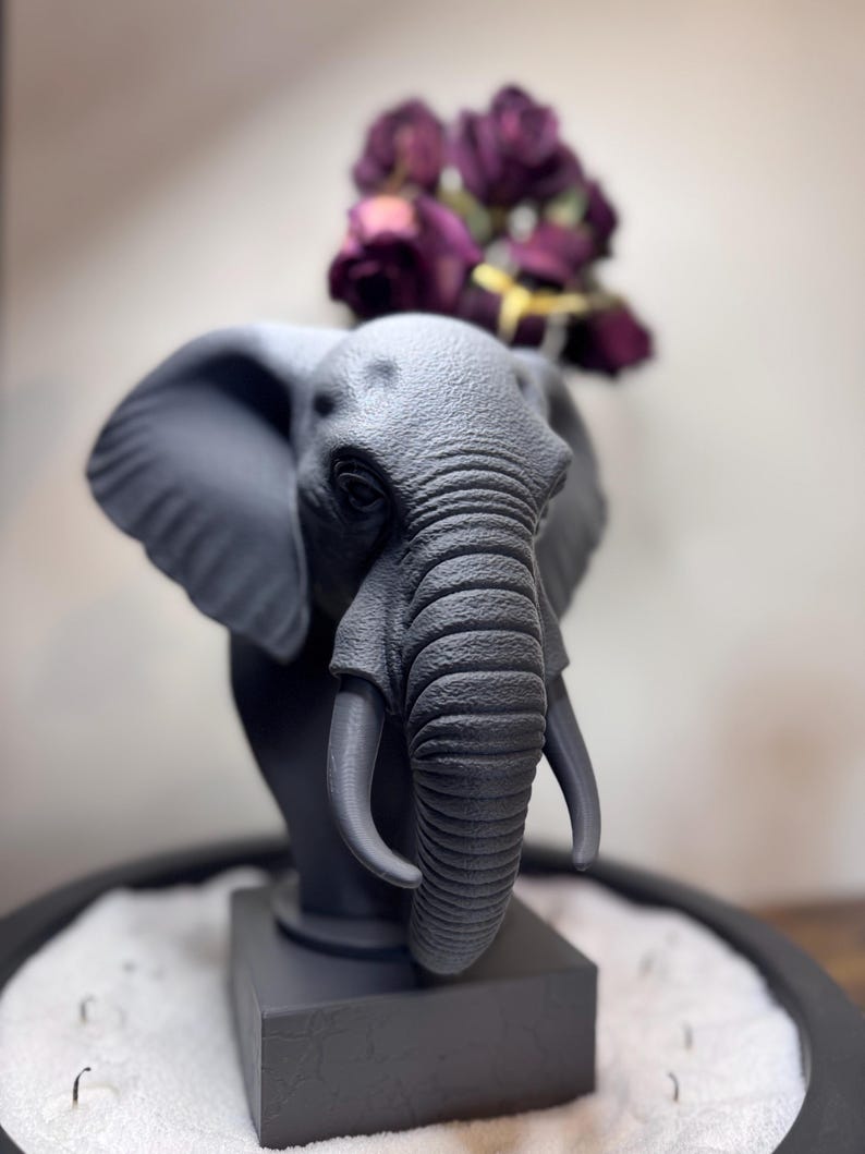 May include: A gray elephant bust with detailed features, including large ears and tusks. The sculpture is mounted on a square base and adorned with a bouquet of deep purple flowers. The overall design is a blend of art and nature.