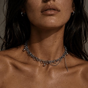 Peut inclure: Un collier argenté composé de plusieurs couches d'anneaux liés et de petites sphères pendantes. Le collier a un design superposé avec une chaîne qui pend. Le bijou est porté autour du cou.