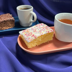 May include: A still life arrangement of cake and drinks. A slice of chocolate cake on a blue tray, with a white mug of coffee. A second cake slice with sprinkles is on a pink tray, with a white mug of tea.