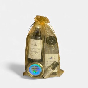 Op de afbeelding: Een verzameling wellnessproducten, waaronder twee flessen, een kleine blik en een roll-on, gepresenteerd in een gouden mesh cadeauzakje. De flessen hebben labels met de tekst "Grandma's Life Syrup" en "Blessed Ground". Het blik is gelabeld "Soothe & Ignite Throat Elixir".