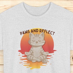 May include: Light grey t-shirt with a cartoon cat meditating in a lotus position against a sunset. The text "PAWS AND REFLECT" is above the cat. The shirt is made of a soft material.