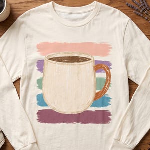 Könnte beinhalten: Ein cremefarbenes Langarmshirt mit einer zentralen Grafik einer beigen Kaffeetasse, die mit dunkler Flüssigkeit gefüllt ist. Die Tasse ist vor einem Hintergrund aus fünf horizontalen, strukturierten Pinselstrichen in Rosa, Grün, Blau und Lila platziert.