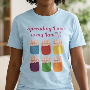 Op de afbeelding: Lichtblauw T-shirt met de tekst "Spreading Love is my Jam" en illustraties van zes kleurrijke jampotten. De potten zijn in tinten rood, geel, groen, paars en oranje. Het shirt is gemaakt van een zacht materiaal.