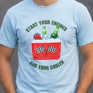 May include: Light blue t-shirt with the text "Start Your Engines and Your Cooler." The graphic features a red cooler with checkered flags, ice, and bottles. The shirt is made of a soft material.