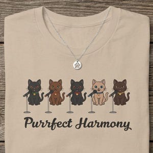 May include: A beige t-shirt featuring a graphic of five cartoon cats standing at microphones with the text "Purrfect Harmony" printed below. A silver-toned necklace with a small paw print pendant rests on the neckline of the shirt.