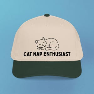 Puede incluir: Una gorra de béisbol de color beige con visera verde oscuro y un botón verde oscuro en la parte superior. El frente presenta un dibujo lineal negro de un gato durmiendo y el texto CAT NAP ENTHUSIAST impreso en una fuente negra, negrita y sans-serif.