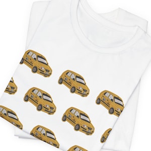Puede incluir: Una camiseta blanca de manga corta con un patrón repetitivo de ilustraciones de taxis amarillos. El estampado gráfico muestra varios coches amarillos pequeños orientados en diversas direcciones sobre la tela de la prenda doblada.