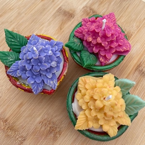 以下が含まれることがあります： 紫、ピンク、黄色の鮮やかな色の3つの花のキャンドル。各キャンドルは、緑の葉が付いたアジサイの花の形をしており、小さな装飾的なボウルに入っています。キャンドルは木製の表面に置かれています。