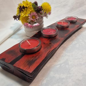Op de afbeelding: Een rood gebeitste houten kandelaar met vier rode kaarsen in glazen. De houder heeft een natuurlijke, golvende rand. Een kleine witte vaas met gele en roze bloemen staat op de achtergrond. Decoratief object.