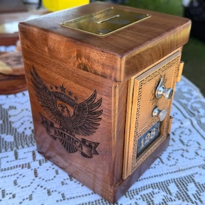 May include: A wooden box with a slot on top and a metal plate. The front features a metal panel with a combination dial and the number 128. An eagle with outstretched wings and the word "AMERICA" is engraved on the side.