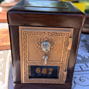 May include: A vintage-style mailbox with a dark brown wooden body and a bronze-colored metal front. The metal front features a star design and the numbers "687" in red. A key hangs from the lock.