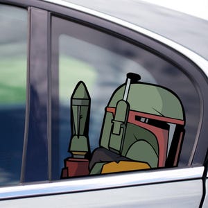 Op de afbeelding: Een autosticker met een cartoonafbeelding van Boba Fetts helm en een raket. De helm is groen, rood en geel, en de raket is groen en bruin. De sticker is op een autoruit geplakt.