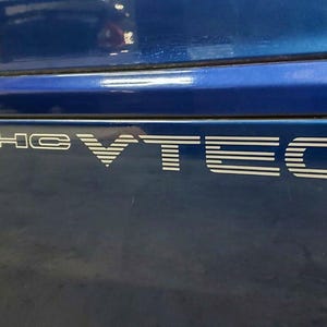 May include: Close-up of a section of a blue car with white lettering. The text reads "DOHC VTEC" in a stylized font. The car's paint has a glossy finish, and the image is well-lit.