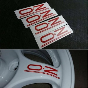 May include: Four white rectangular stickers with the letters "O.Z" in red, with a red line underneath. The bottom image shows a white car wheel with the letters "O.Z" in red.