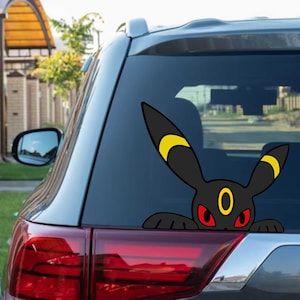 May include: A black car window decal featuring a cartoon Umbreon character. The decal shows the character's head and arms, with red eyes, yellow accents, and a yellow ring. The decal is applied to the rear window of a car.