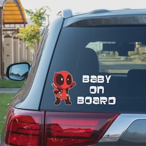 Może przedstawiać: Naklejka na szybę samochodową z czerwoną i czarną postacią z kreskówki Deadpool trzymającą broń. Biały tekst brzmi "BABY ON BOARD", wskazując na obecność dziecka w pojeździe.