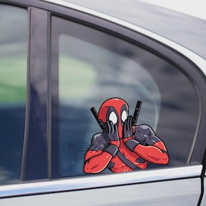 Op de afbeelding: Een rood-zwarte Deadpool auto sticker. Het stripfiguur houdt twee zwarte pistolen vast en bedekt zijn ogen met zijn handen. De sticker is aangebracht op een autoruit.