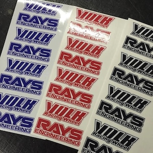 May include: Sticker sheets displaying the "VOLK Racing Wheel" and "RAYS ENGINEERING" logos in blue, red, and black. The stickers have a shiny surface and are arranged in rows.