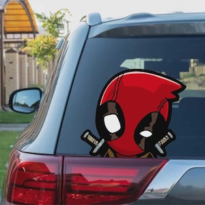 Może przedstawiać: Czerwono-czarna naklejka na samochód Deadpool. Naklejka przedstawia kreskówkową wersję Deadpoola z dwoma mieczami. Naklejka jest przymocowana do tylnej szyby szarego samochodu.