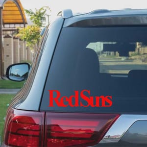 May include: A red "Red Suns" vinyl sticker is applied to the rear window of a grey car. The text is in a bold, sans-serif font. The car's rear light is visible below the sticker, with a glimpse of the side mirror.