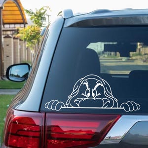 Pode incluir: Autocolante de vinil branco com o rosto de uma personagem de desenho animado, espreitando por cima de uma linha horizontal. O autocolante está aplicado no vidro traseiro de um carro cinzento.