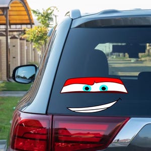 May include: A car window decal featuring a cartoon face with a red and white top, blue eyes, and a smiling mouth. The decal is applied to the rear window of a gray SUV, with a blurred background of a residential area.