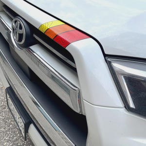 Puede incluir: Primer plano de la parrilla delantera de un coche blanco con una franja horizontal colorida. La franja pasa de amarillo a naranja a rojo, con líneas de contorno negras. El emblema de Toyota es visible.