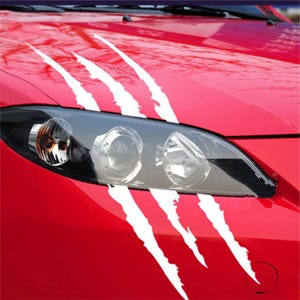 May include: Red car bonnet with white claw mark decals. The decals are angled and appear to be tearing through the bonnet. The car's headlight is visible below the decals.