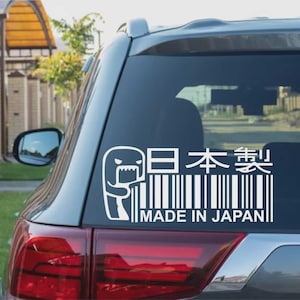 May include: White vinyl decal on a car window featuring a cartoon character, a barcode, and Japanese text. The text reads "Made in Japan" and "日本製". The decal is applied to the rear window of a gray vehicle.