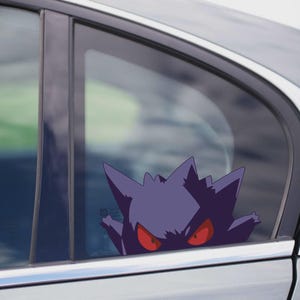 May include: A purple Gengar car sticker with red eyes is applied to a car window. The sticker is a stylized illustration of the Pokémon character, appearing from behind the window frame. The car is silver.