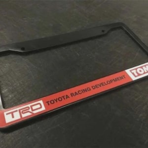 May include: Black license plate frame with a red and white accent featuring the text "TOYOTA RACING DEVELOPMENT" and the TRD logo. The frame also includes the "TOM'S" logo.