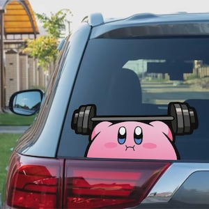 May include: A pink cartoon character sticker on a car window, holding a barbell. The character has large blue eyes and a determined expression. The barbell is black and grey, and the sticker is placed on the rear window of a vehicle.