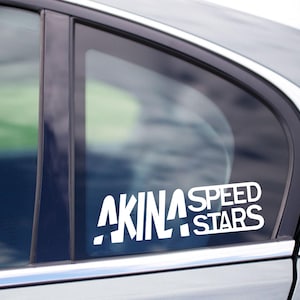 Peut inclure: Autocollant de voiture en vinyle blanc avec le texte "AKINA SPEED STARS" dans une police de caractères audacieuse et stylisée. L'autocollant est apposé sur la vitre d'une voiture argentée, le texte étant bien visible.