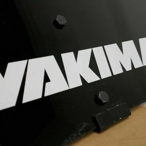 May include: Close-up of a black metal panel featuring the white word "YAKIMA". The logo is in a bold, sans-serif font. The panel is secured with black bolts, suggesting a product component.
