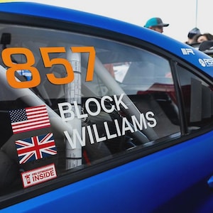 May include: A blue race car window with the number 857 in orange. The name "BLOCK WILLIAMS" is in white text. American and British flags are displayed, along with a "FIRE EXTINGUISHER INSIDE" sticker.
