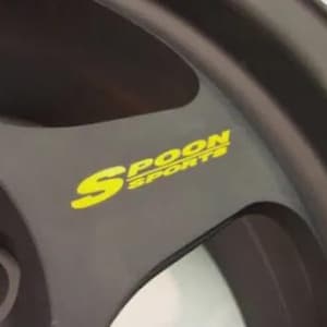 Może przedstawiać: Zbliżenie czarnego koła samochodowego z żółtym napisem "SPOON SPORTS". Koło ma ciemnoszare wykończenie i wieloramienną konstrukcję. Obraz jest dobrze oświetlony, podkreślając szczegóły koła.