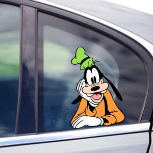 Può includere: Un adesivo in vinile di Pippo, un personaggio dei cartoni animati Disney, è applicato sul finestrino di un'auto. Pippo è raffigurato con il suo caratteristico cappello verde, la camicia arancione e i guanti bianchi.
