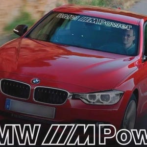 Puede incluir: Un sedán BMW rojo con el texto "BMW ///M Power" en el parabrisas y en la parte inferior de la imagen. El coche está en movimiento en una carretera, con un conductor visible en el interior.