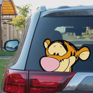 Peut inclure: Autocollant en vinyle coloré représentant Tigrou de Winnie l'ourson. L'autocollant, de couleur orange et noire avec un nez rose, est appliqué sur la vitre arrière d'une voiture. Le personnage de dessin animé regarde par-dessus le bord.