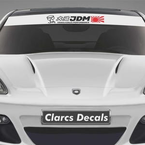 May include: White JDM banner decal with black and red graphics, including Japanese text and a rising sun design. The decal is displayed on the windscreen of a white car. The text "Clarcs Decals" is displayed on the front bumper.